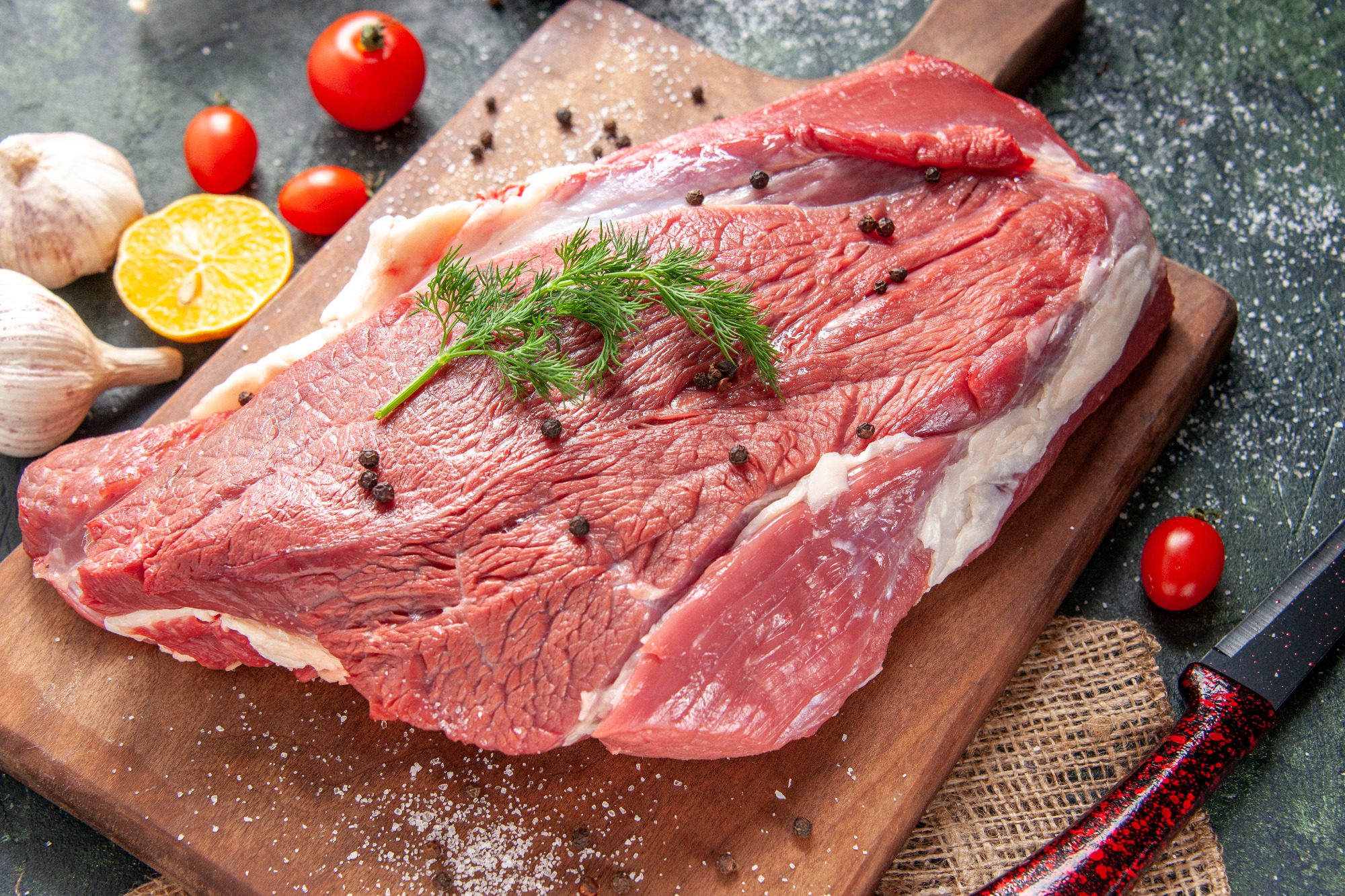 Rohes Fleischstück auf einem Holzbrett, umgeben von Gewürzen und Tomaten.
