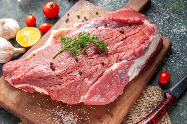 Rohes Fleischstück auf einem Holzbrett, umgeben von Gewürzen und frischen Tomaten.